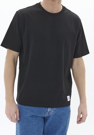Černé tričko s kulatým výstřihem z hladké látky, s krátkými rukávy a malou bílou štítkem na lemu. Kombinováno s modrými džíny.