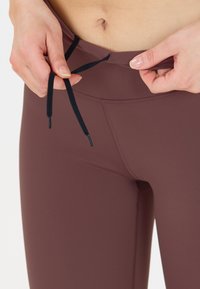 Bruna leggings gjorda av elastiskt tyg, med en svart dragsko i midjan. Närbild visar midjebandet och sömnadsdetaljer.