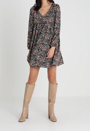 Kobieta w długim, kwiecistym płaszczu z dekoltem w kształcie litery V, w połączeniu z beżowymi kozaki na obcasie sięgającymi do kolan, na jednolitym tle.