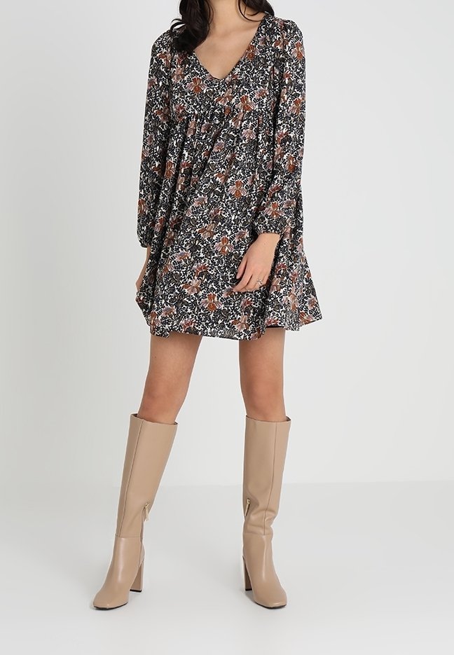 Femme portant une robe fleurie à manches longues avec un col en V, associée à des bottes beige à talons montant jusqu'au genou, sur un fond uni.