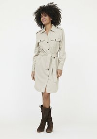 Beiges Hemdkleid mit langen Ärmeln und Knopfleiste, ausgestattet mit zwei vorderen Taschen und einem gebundenen Taillengürtel, kombiniert mit braunen Stiefeln bis zum Knie.