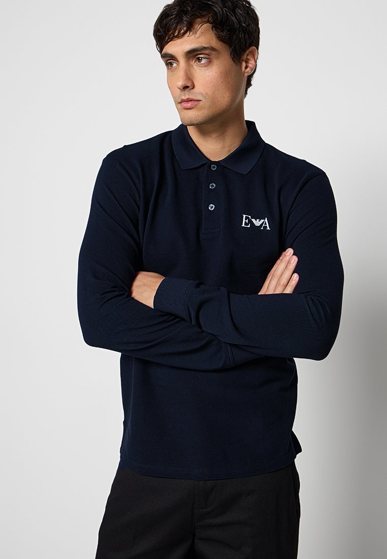 Hombre con los brazos cruzados, vestido con un polo de manga larga azul marino con el logo blanco "EVA" en el pecho, mirando hacia un lado.
