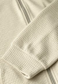 Chaqueta color crema con un tejido texturizado de punto gofrado. Presenta una cremallera plateada y puños acanalados. La superficie texturizada realza el detalle visual.