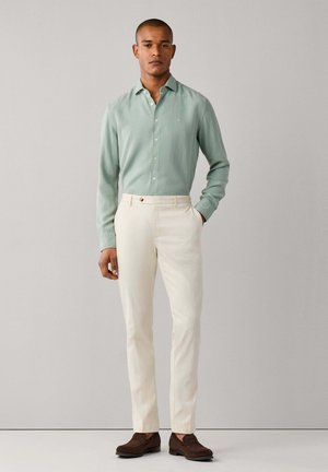 Hombre de pie frente a un fondo liso con camisa de botones verde claro, pantalones crema y zapatos de cuero marrón oscuro.