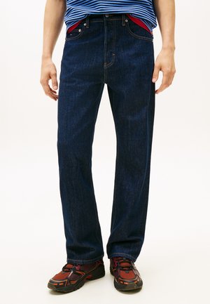 Personne portant un jean droit bleu foncé, avec une couche rouge visible à la taille, une chemise rayée bleue et des baskets noires et rouges.