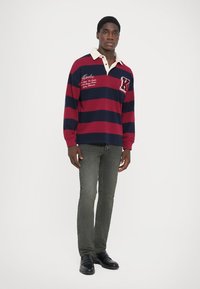 Rood en marineblauw gestreepte lange mouwen polo met een witte kraag en logo-detail, gecombineerd met slanke olijfgroene jeans en zwarte schoenen.