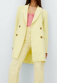 Double-breasted lichtgele blazer met gouden knopen; heeft een getailleerd ontwerp en inkeping revers, gecombineerd met een pastelroze top en gele broek.