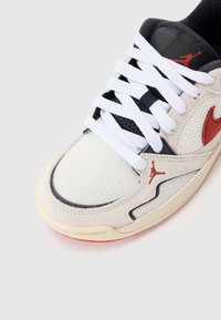 Białe siatkowe i zamszowe sneakersy z czarnymi akcentami i czerwonym logo. Posiadają płaskie białe sznurówki i zakrzywiony fason. Gumowa podeszwa z delikatną wariacją kolorystyczną.
