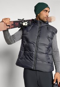 Granatowa kamizelka puchowa z wysokim kołnierzem, zamkiem błyskawicznym i pikowanym wzorem. Model trzyma narty i nosi zieloną czapkę oraz szary top z długim rękawem.