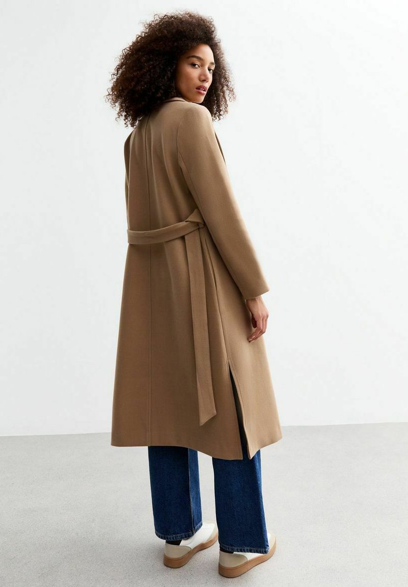 Cappotto lungo color cammello con cintura, ventaglio posteriore e taglio dritto, abbinato a jeans a gamba larga blu e sneakers bianche. Texture liscia.