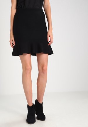 Femme portant une jupe noire ajustée avec un ourlet évasé et des bottines noires, debout devant un fond clair uni.