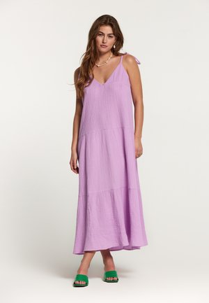 Lila, Maxikleid mit V-Ausschnitt, strukturiertem Stoff und Spaghetti-Trägern. Verfügt über ein gestuftes Design. Getragen mit grünen Slidesandalen.