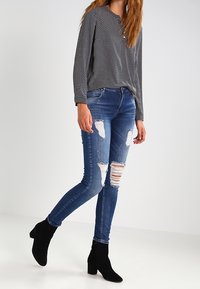 Jean bleu délavé et usé avec une coupe slim, présentant plusieurs déchirures et un effet délavé. Associé à un chemisier à manches longues noir à pois blancs et des bottines noires.