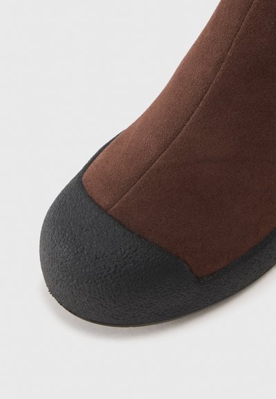 Bottine en daim marron avec un embout en caoutchouc noir texturé. Tige lisse avec un design élégant et ajusté.