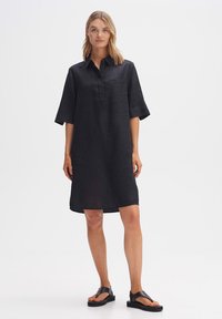 Abito camicia in lino nero con colletto, maniche corte, una tasca frontale e una silhouette dritta, abbinato a sandali piatti neri.