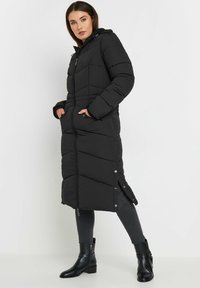 Cappotto puffer nero con cappuccio, lunghezza lunga, texture trapuntata, tasche laterali e chiusura con zip. Abbinato a stivaletti neri alla caviglia e jeans skinny.