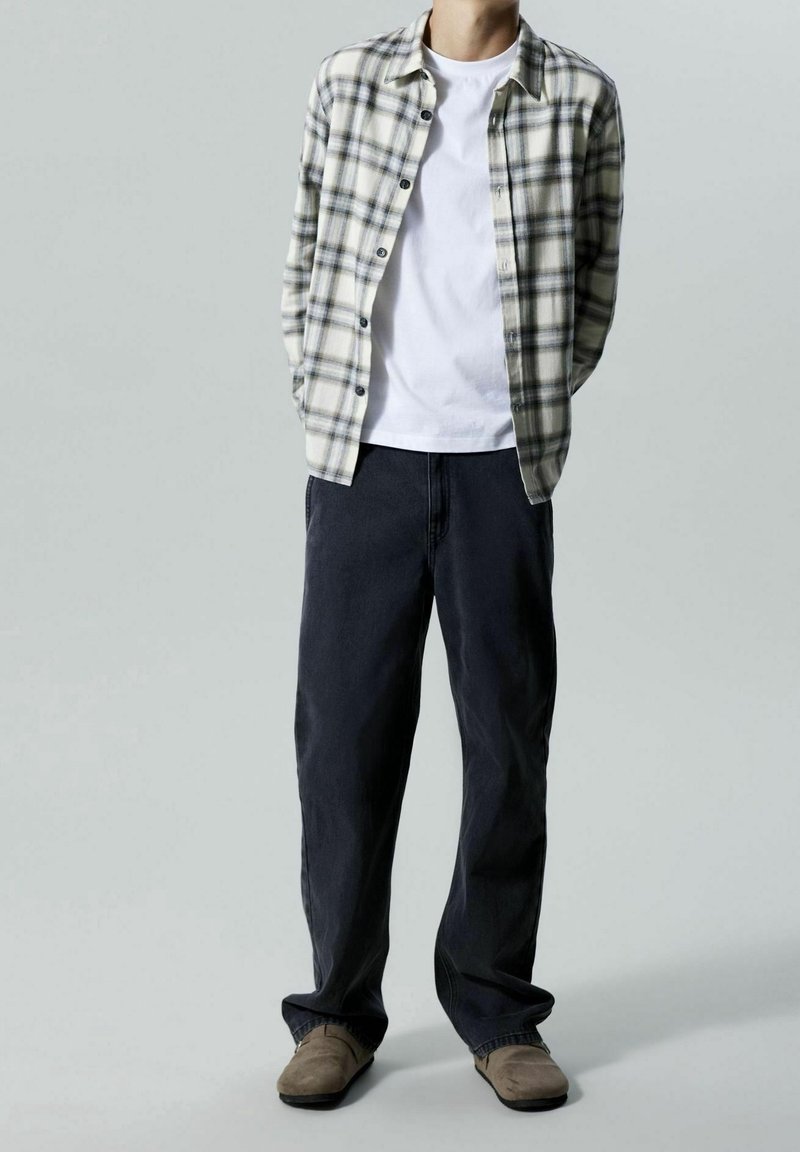 Homme portant un pantalon noir, des chaussures beiges, un t-shirt blanc et une chemise à carreaux ouverte les mains dans le dos sur fond gris.