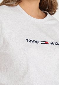 T-shirt in cotone grigio chiaro con scollo rotondo. Presenta la scritta "TOMMY" e "JEANS" in ricamo blu navy e rosso, con un design del logo orizzontale.