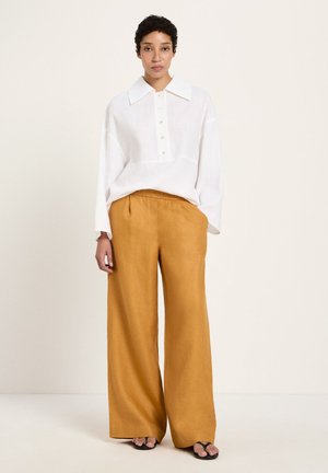 Person wearing a loose white long-sleeve shirt and wide mustard-colored pants standing against a plain light background.