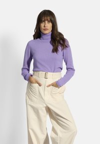 Lila Rollkragenpullover aus weichem Stoff mit langen Ärmeln. Kombiniert mit hochgeschnittenen, weit geschnittenen Sahne-Hosen mit großen Taschen.