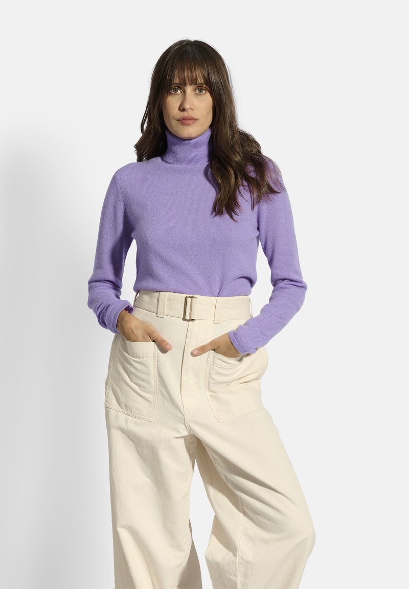 Lila Rollkragenpullover aus weichem Stoff mit langen Ärmeln. Kombiniert mit hochgeschnittenen, weit geschnittenen Sahne-Hosen mit großen Taschen.