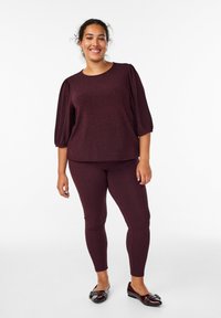 Burgundin värinen teksturoitu paita puffihihnoilla, yhdistettynä samansävyisiin leggingseihin ja tummiin tasapohjaisiin kenkiin. Asussa on hienovarainen herringbone-kuvio.