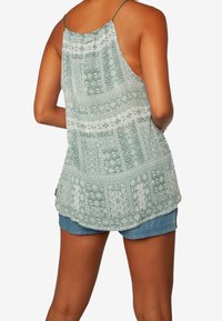 Femme portant un haut vert à motifs sans manches avec de fines bretelles et un short en jean bleu, vue de dos sur un fond blanc.