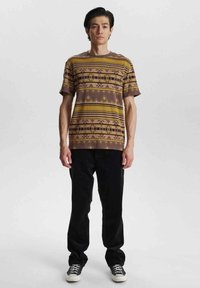 Camisa de manga corta marrón con patrones geométricos amarillos, combinada con pantalones negros y zapatillas negras. Diseño simple y ajuste relajado.