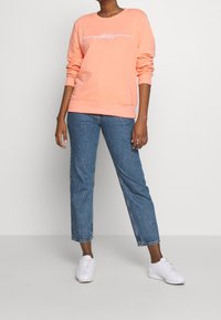 Koraal sweatshirt met witte scriptlogo, gecombineerd met blauwe denim jeans met bloembroderie, gedragen met witte sneakers.