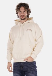 Felpa color crema con tasca frontale, cappuccio regolabile, e testo "PROPAGANDA" in un font curvo sul lato sinistro del petto. Texture morbida.