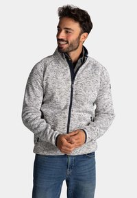 Man die glimlacht en naar links kijkt, gekleed in een lichtgrijze fleecejack met rits en een patroon, en blauwe spijkerbroek, met de handen losjes voor zich geklemd.