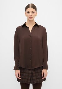 Camisa de botón de color marrón oscuro con mangas largas, textura suave y cuello. Cuenta con botones a juego en la parte frontal. Estilizada con una falda de cuadros.