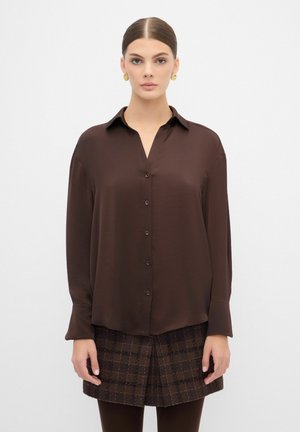 Chemise boutonnée marron foncé à manches longues, texture lisse et col. Présente des boutons assortis le long de l'avant. Associée à une jupe à carreaux.