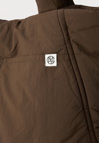 Giacca isolante marrone con finitura testurizzata, caratterizzata da un'etichetta bianca con un logo cucito nell'angolo superiore sinistro della tasca.