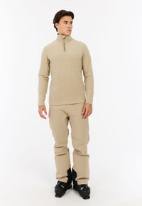 Lys beige thermotop med høj hals, halv lynlås og lange ærmer, parret med beige snebukser og sorte skistøvler.