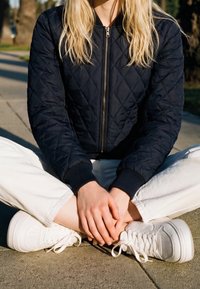 Personne portant une veste matelassée noire, un pantalon blanc et des baskets blanches assise jambes croisées sur un trottoir avec les mains posées sur les pieds.