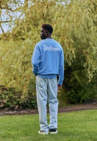 Mees seisab murul, näoga suurte rohelise puu poole, kandes helerohelist dressipluusi, millel on tekst "Soulscape", ning helerohelisi teksapükse ja tossusid.