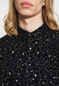 Černá košile s leopardím potiskem, zdobená zlatými tečkami. Vyrobena z jemné tkaniny, má klasický límeček a zapínání na knoflíky.