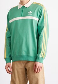 Grön sweatshirt med vit rand, dragkedja vid kragen och tre gula ränder på varje ärm. Har adidas-logga på bröstet.