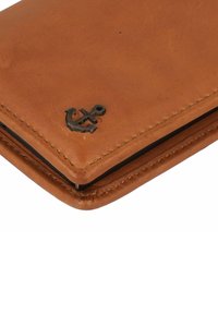 Bruine leren portemonnee met gestikte randen, voorzien van een metalen ankerembleem in de onderste hoek en een gladde textuur.