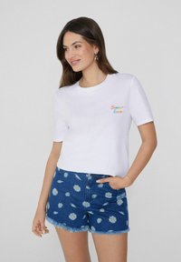 Camiseta de algodón blanca con "Verano Amante" bordado en texto colorido, combinada con pantalones cortos de mezclilla azules con un estampado de margaritas y dobladillo desflecado.