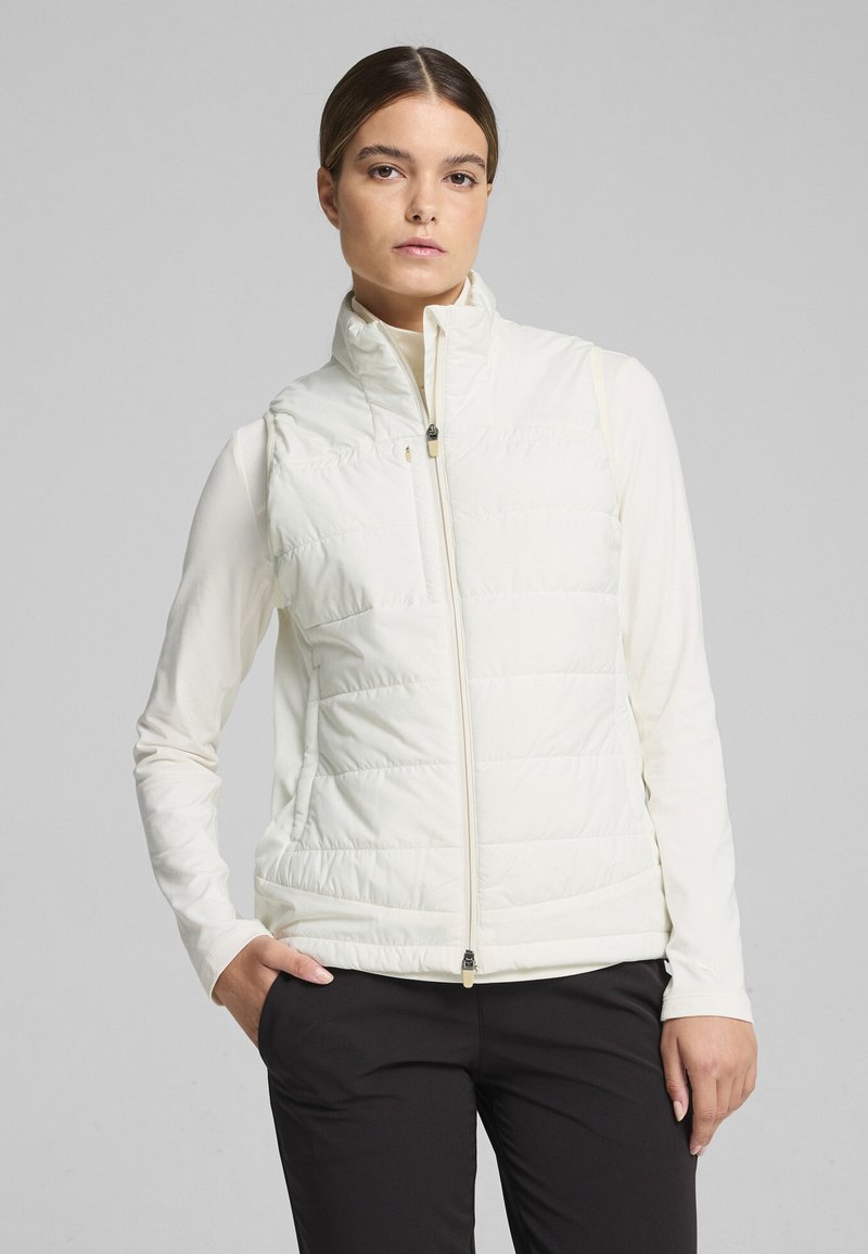 White padded vest with a high collar, front zipper, and side pockets, worn over a long-sleeve top. Smooth texture with a quilted design.