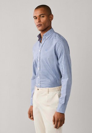 Ein Mann trägt ein blau kariertes Hemd mit Knöpfen, das in beige, hoch taillierte Hosen gesteckt ist, vor einem schlichten Hintergrund.