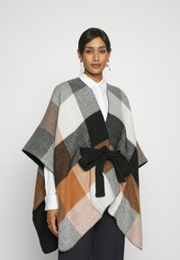 Poncho de punto en cuadros negro, gris, blanco y marrón, con un cinturón de tela negra. Presenta un diseño holgado y una textura suave.