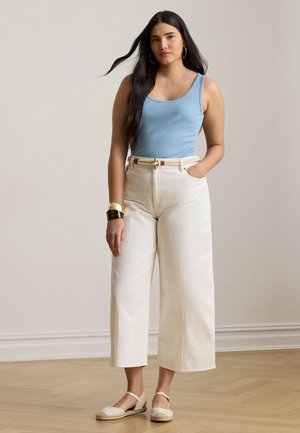 Femme aux longs cheveux noirs portant un haut sans manches bleu clair, un pantalon blanc à jambes larges avec une ceinture en corde, des bracelets dorés et bruns, et des ballerines beiges.