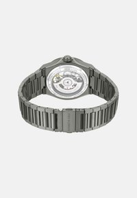 Roestvrijstalen horloge met een slanke, achthoekige kast en een gepolijste grijze band. De transparante kast achter toont de beweging en merkdetails.