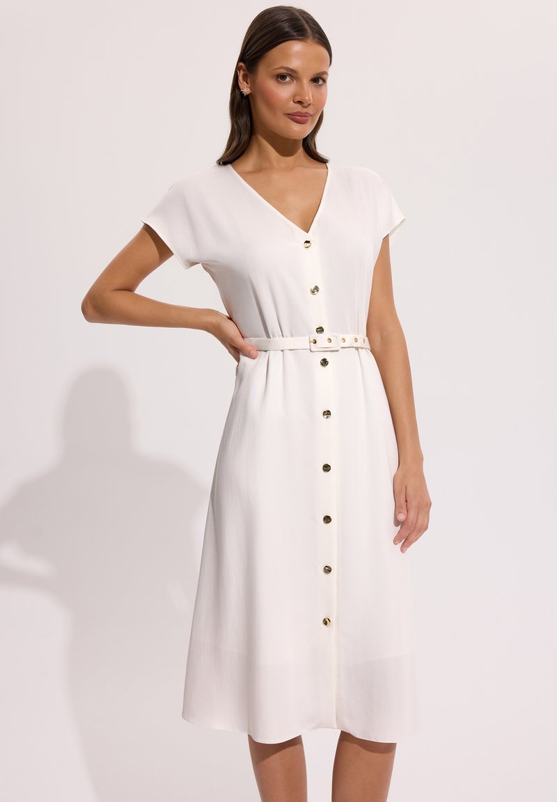 Kobieta ubrana w białą sukienkę zapinaną na guziki do kolan, z dekoltem w serek i pasem w tym samym kolorze, stojąca z jedną ręką na biodrze.