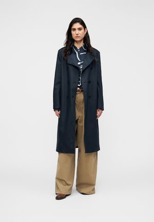 Granatowy długi płaszcz typu trencz z dwurzędowym zapięciem, szerokimi klapami i czarnymi guzikami; w połączeniu z szerokimi khaki spodniami i pasiastą koszulą.