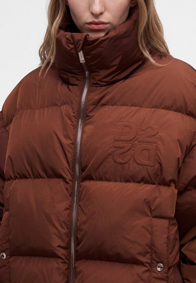 Femme portant une veste matelassée marron à col haut zippé, avec le logo "DS2" brodé sur la poitrine.