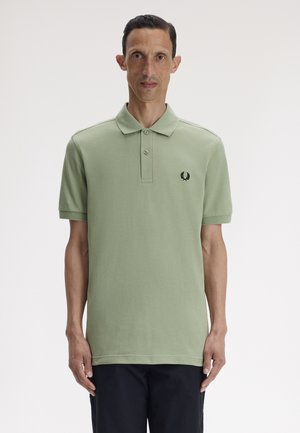 Mand står fremadvendt iført en lysegrøn kortærmet poloskjorte med logo på brystet og mørke bukser mod en ensfarvet baggrund.